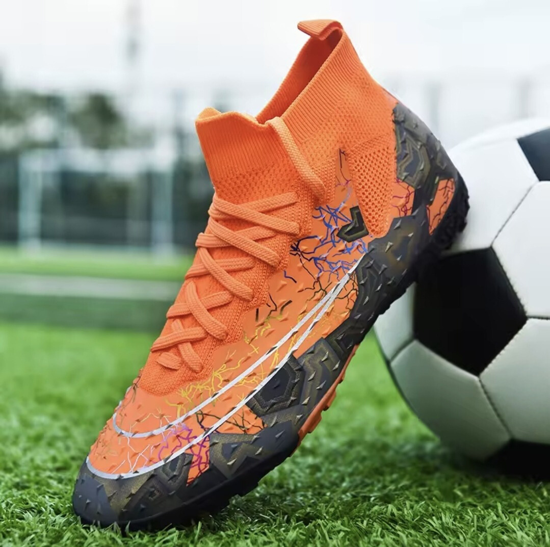 Tempesta Men’s Firm Ground Football Boots in orange with black sole and colourful crack details shown on a football pitch next to a ball