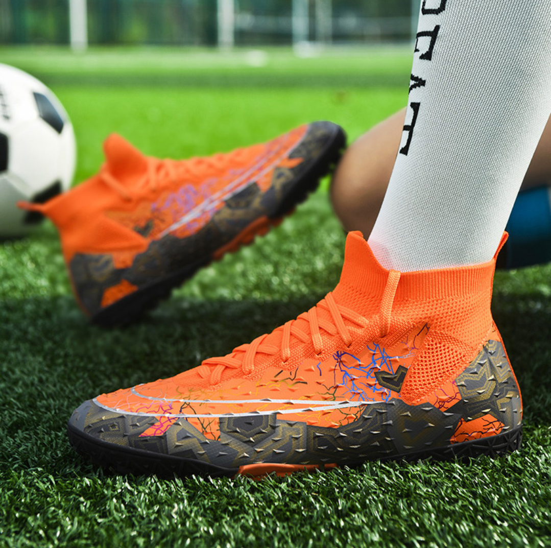 Tempesta Men’s Turf Football Boots in orange with black graphic details worn on artificial grass field with a football in the background
