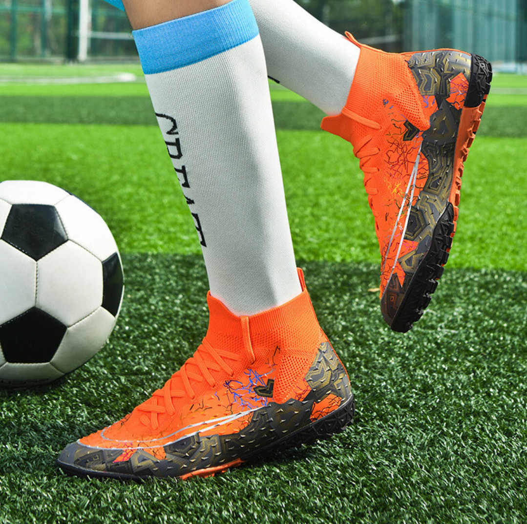 Tempesta Men’s Turf Football Boots in orange with black graphic details worn on artificial grass field with a football in the background