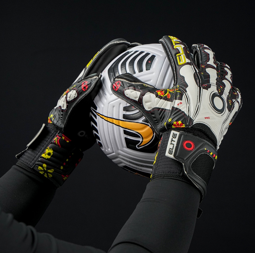 Goalkeeper holding football with Elite Sport Pro Calavera gloves featuring skeleton and floral details on black background