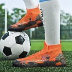 Tempesta Men’s Turf Football Boots in orange with black graphic details worn on artificial grass field with a football in the background