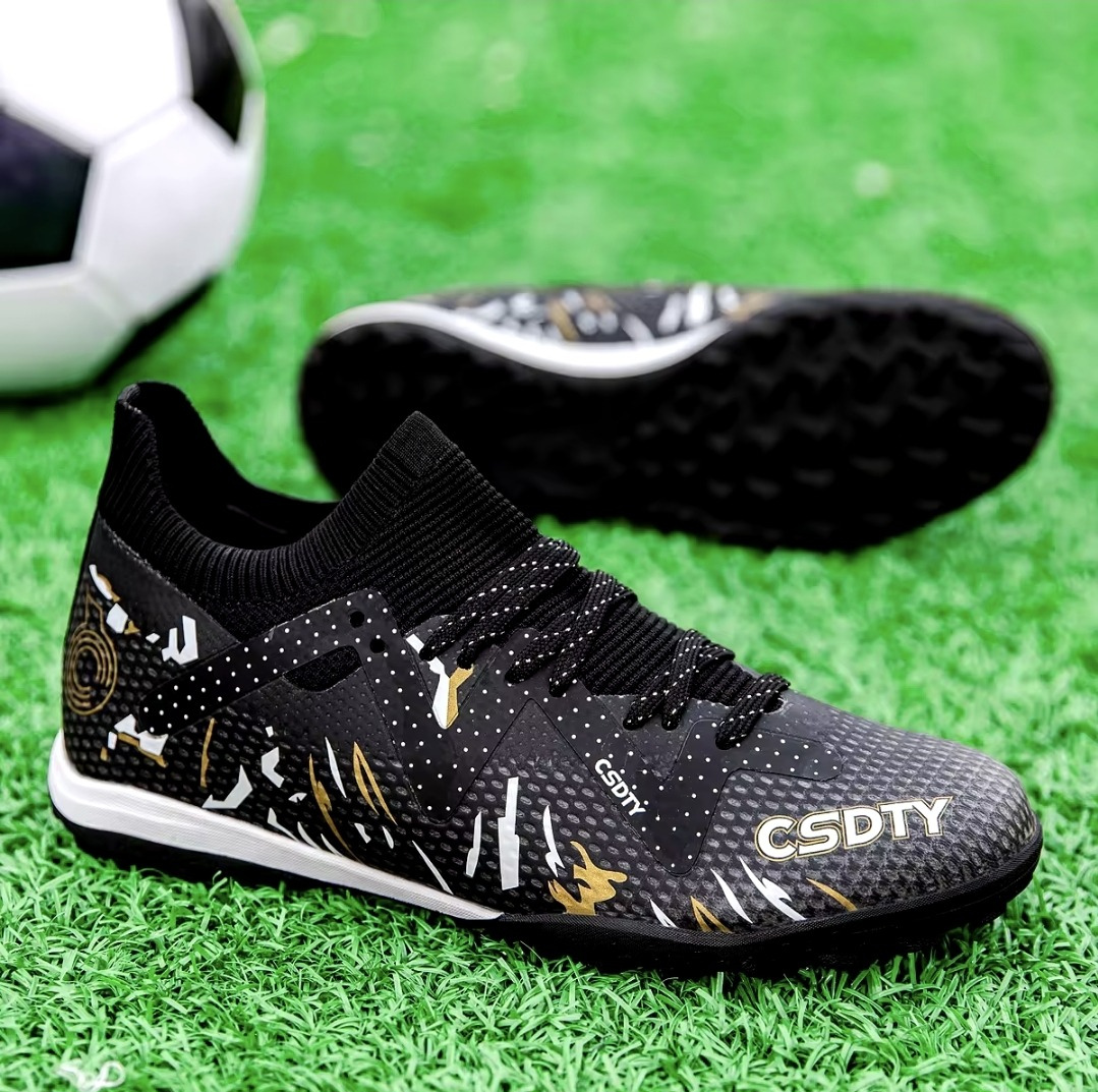 Pair of Galactic Men’s Turf Football Boots black patterned next to a football on green artificial grass field background