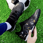 Galactic Men’s Turf Football Boot black patterned on foot next to a football on green artificial grass field background