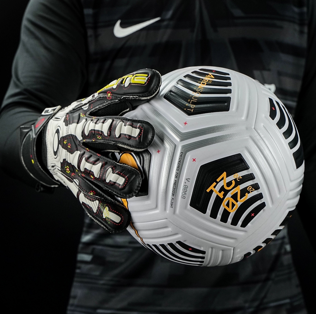 Goalkeeper holding football with Elite Sport Pro Calavera gloves featuring skeleton and floral details on black background