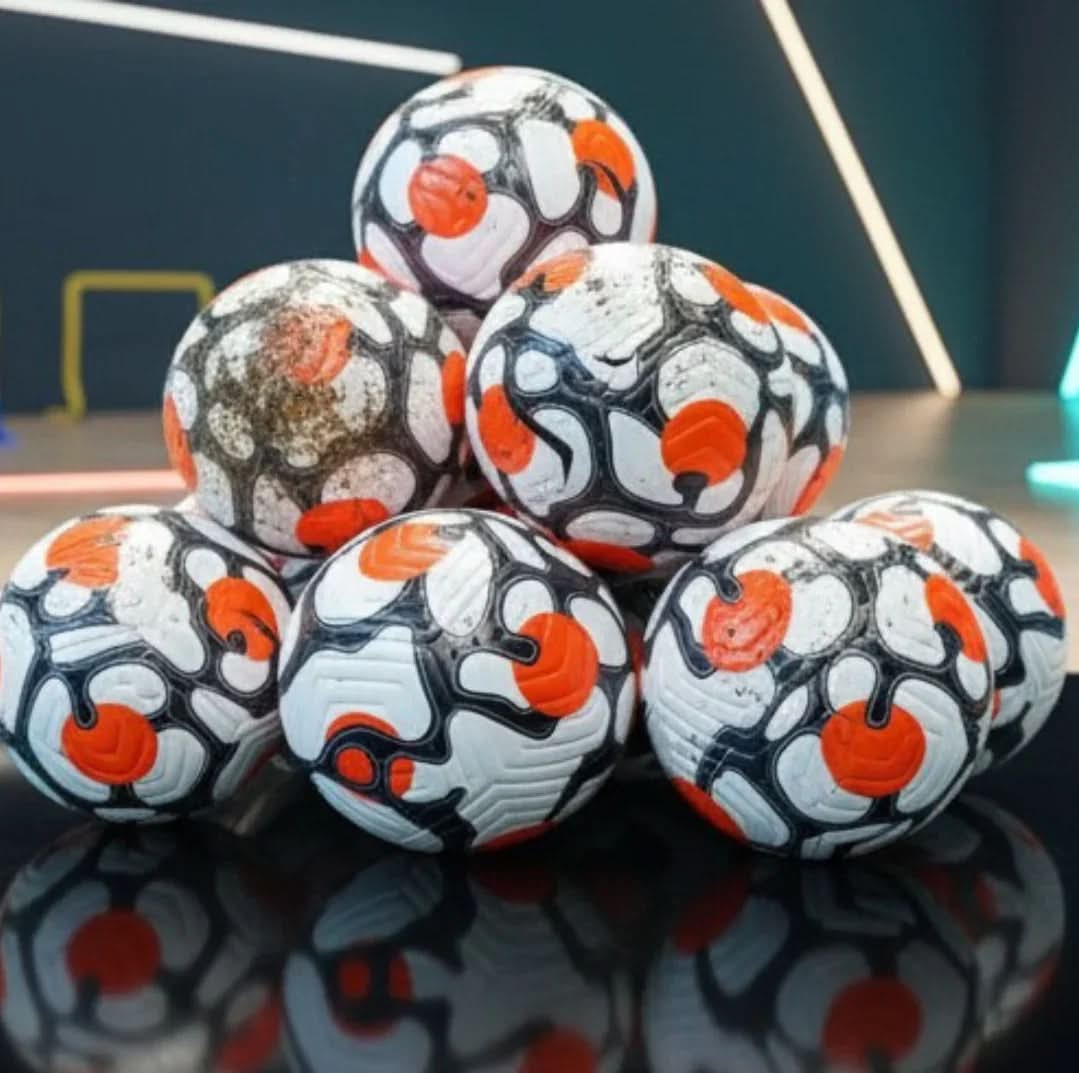 A pyramid stack of seven footballs, featuring a white, black, and orange design, resting on a shiny black surface. One ball in the centre-left is noticeably covered in mud. The background is a blurred, dark blue room with neon light accents.