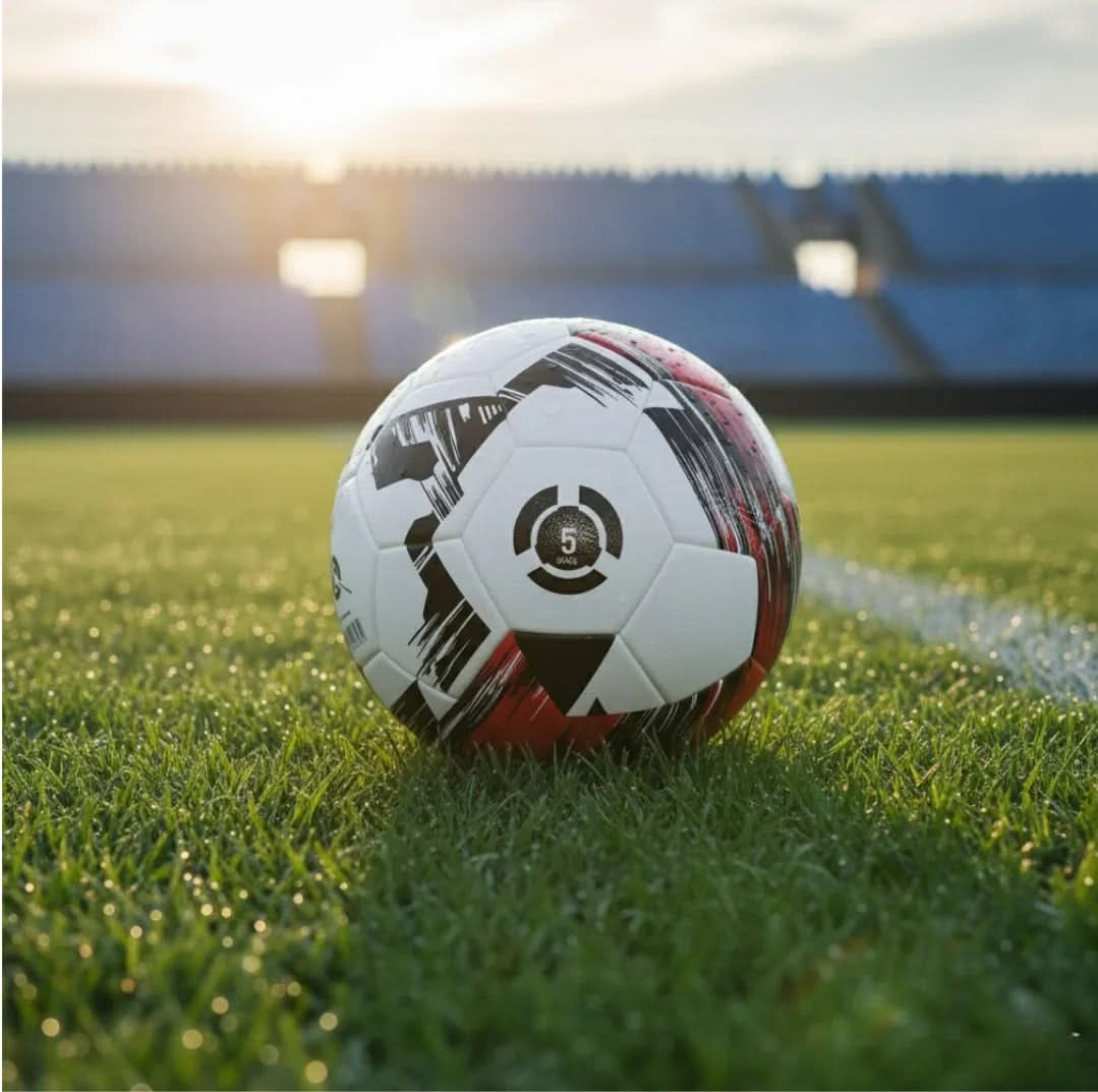 Football resting on a green pitch with stadium seats in the background, sunlight shining.